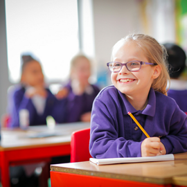 Primary aged girl wearing purple uniform looks off into the distance smiling with a pencil in her hand, actively engaging in classroom activites.