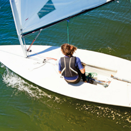 Lady sailing a white boat with a white sail. The image is taken from above looking down on the back of the lady. She is wearing a buoyancy aid and a purple t-shirt.