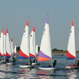 Approx 10 sailing dinghy's being sailed by children, in a group together. They are on a lake with blue sky. The sails are mainly white with red and blue or orange and purple flashes of colour on.