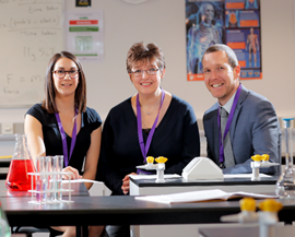 Three smiling individuals wearing purple lanyards sit at a science lab table with test tubes and a red liquid-filled flask. Educational posters are on the wall.