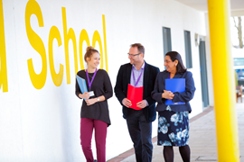 three people, holding files, walking along a corridor with a wall with School in yellow written on it.