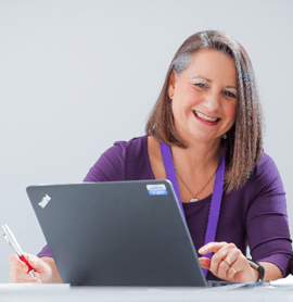 Lady sitting at desk behind laptop. Laptop is open and she is looking at the screen. She is wearing a purple top and is smiling.