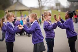 Four primary aged girls  in purple sweaters are playing a game, clapping hands in pairs on a school playground, surrounded by other kids.