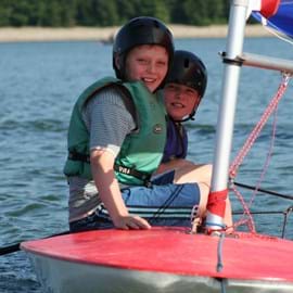 Two boys wearing buoyancy aids and black hard hats sailing in the water on a red dinghy type boat. Both boys are smiling and having fun.