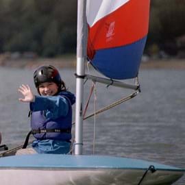 Girl wearing black hard hat and life jacket sailing a blue dingy with a red, white and blue sail blowing in the wind