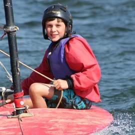 Primary aged boy, wearing protective clothing, a life jacket and a black hard hat, sailing a red boat, smiling