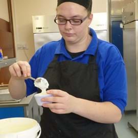 A person in a blue shirt and black apron is carefully spooning white dessert into a small cup in a kitchen.