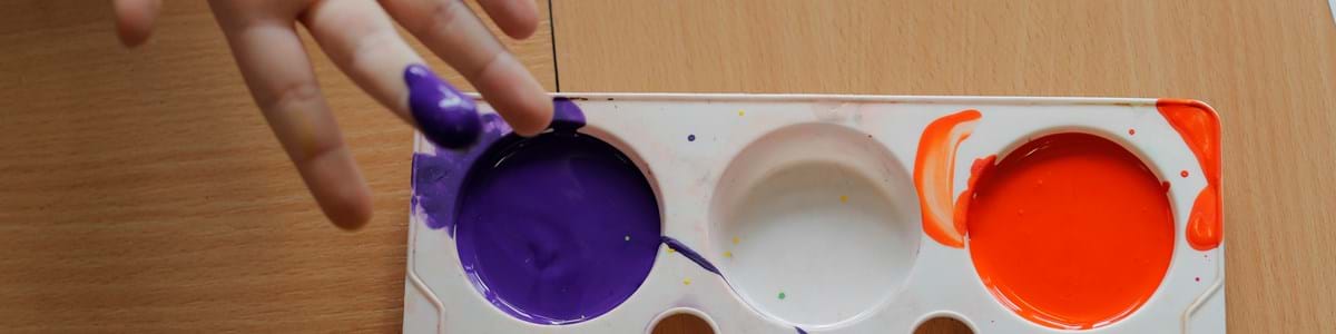 A hand with a purple-painted fingertip reaches toward a paint tray. The tray holds wet purple and orange paint, set on a wooden surface