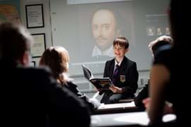 A smiling boy in a school uniform reads Romeo and Juliet aloud to classmates in a classroom. A portrait of Shakespeare is projected on the screen behind him.