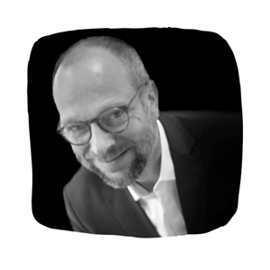 A smiling man with glasses and a beard is in a suit and tie against a black background. 