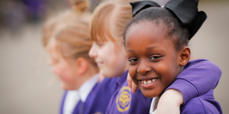A young primary aged girl smiles at the camera whilst embracing her friends.