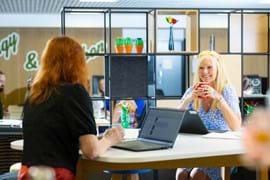 Two women in a modern office, one with red hair sits with a laptop, back to camera, while the other in a floral dress smiles, holding a red mug.