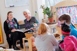 Two women with purple lanyards sit in a well-lit early childhood education setting, engaged in discussion with notebooks in hand. In the foreground, a group of children interact with learning materials, while a wooden shelf with baskets, plants, and various educational items adds warmth to the environment. A colorful curtain and a whiteboard with notes are visible in the background.