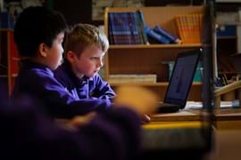 Two young boy students in purple uniforms focus intently on a laptop in a classroom filled with books.