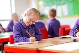 Primary aged girl in a classroom, sitting at a desk, writing down on paper with a pencil.
