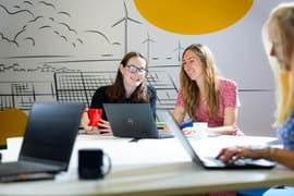 Two women are sitting at a bright, modern table in a collaborative workspace, looking at a laptop together and smiling One holds a red coffee mug, while the other, wearing a pink dress, appears engaged in conversation. The workspace features laptops, coffee cups, and a creative wall mural with solar panels and wind turbines