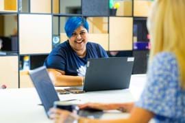 A woman with blue hair, wearing a navy blue t-shirt, smiles while sitting at a table with a laptop in front of her. Another person, slightly out of focus, works on a laptop in the foreground. The room features modern shelving and a collaborative workspace atmosphere.