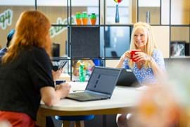 Two women sit at a table in a modern office, smiling and engaged in conversation. One holds a red mug while another works on a laptop. The space features shelves with colorful decor, creating a bright, collaborative atmosphere.