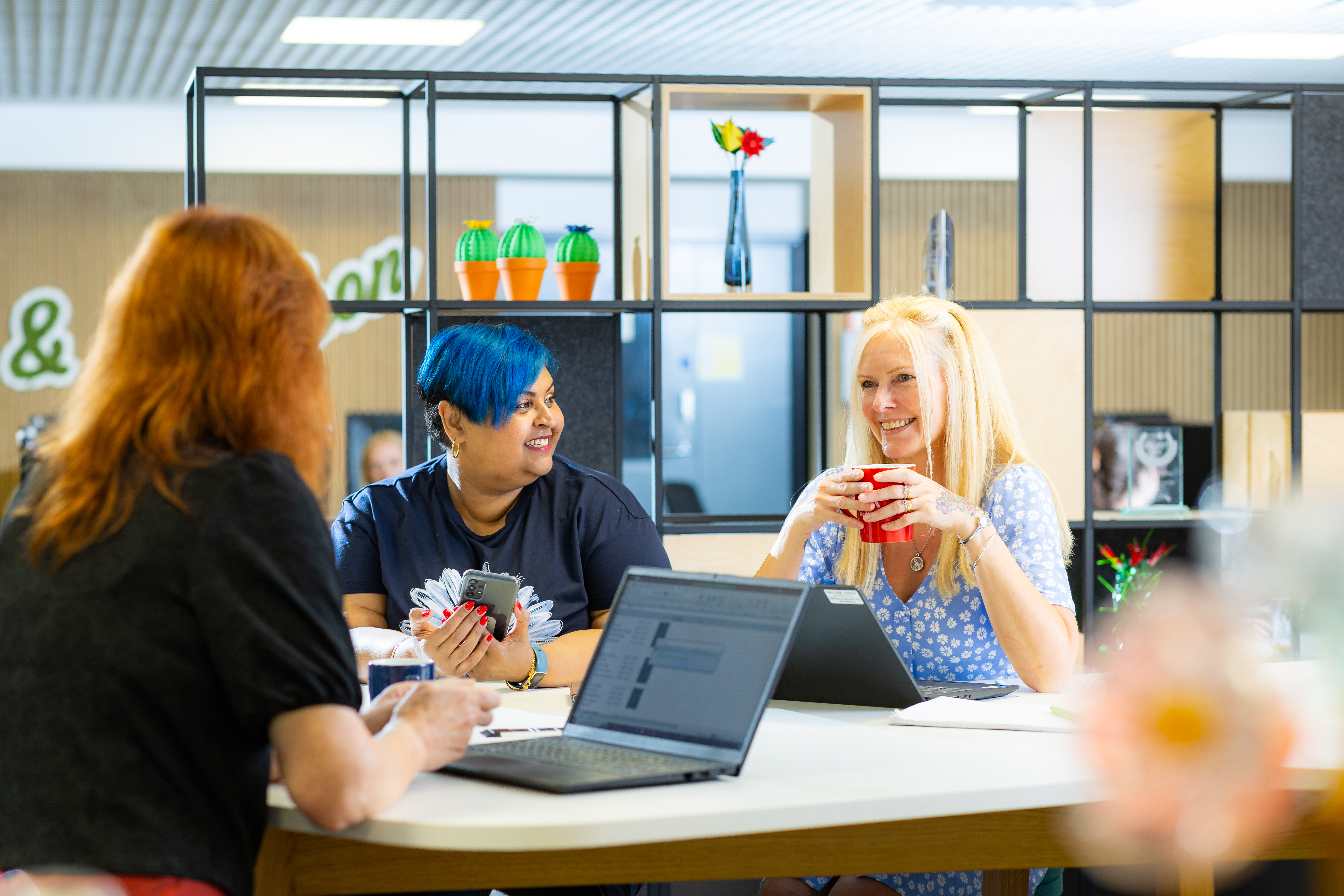 Three female colleagues sitting at a table with laptops, talking and holding papers. Shelving with decor is in the background.