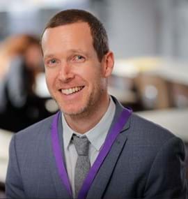 A smiling man wearing a grey suit and a purple lanyard, with a light grey shirt and a grey tie, is seated in a school setting.
