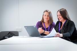 Two women with brown hair sit side by side at a desk, both wearing purple lanyards. They are focused on a laptop screen in front of them, appearing engaged in discussion or collaboration. The desk is plain, and the background is neutral.