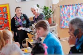 A nursery setting with two educators seated in the background, one holding a book, surrounded by colourful decorations, plants, and toys. Children and a staff member are visible in the foreground, including a woman on the phone.