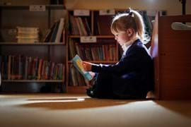 A young girl with blonde hair sits cross-legged on the floor in a library, reading a book with focus. Bookshelves filled with colorful books line the background, while soft lighting highlights her and creates a warm, quiet atmosphere.