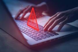 The image shows a pair of hands typing on a laptop keyboard with a glowing red warning triangle and exclamation mark superimposed. The scene conveys a sense of urgency or cyber security concerns, highlighting risks such as hacking, data breaches, or system vulnerabilities.