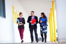 Three educators walking together outside a school building, engaged in conversation and holding colourful folders. The area is supported by bright yellow pillars