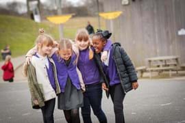 Four young primary school girls wearing school uniforms walking together outdoors, laughing and holding each other closely, symbolizing friendship and joy. A playground with a picnic table and green grass is visible in the background.