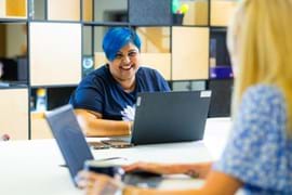 Smiling person with blue hair and a blue shirt works on a laptop at a table with another person.