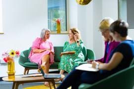 Four women engage in a lively discussion on green couches. One wears a pink dress, another a green floral dress. Two more are blurred in the foreground.