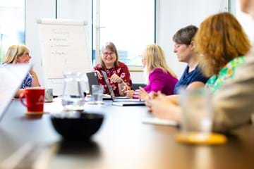 A group of people sitting round a large desk undertaking a training session, they are looking towards the front to the trainer who is smiling at them all.