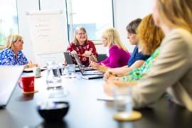 A diverse group of five people sit around a conference table in a bright room, engaging in lively discussion. A flipchart lists training options.