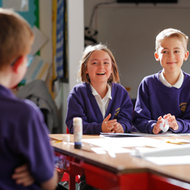 Three primary school children in purple uniforms sit around a classroom table, engaged in a lively discussion. Two of them are laughing while the third listens attentively.