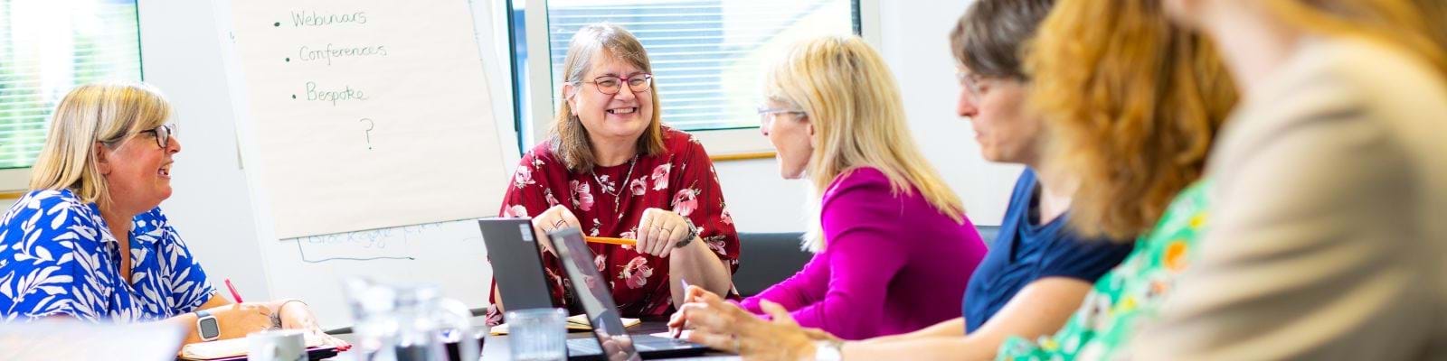 A group of women sit at a conference table, engaged in discussion. One woman in a red floral shirt smiles while holding a pencil, creating a positive atmosphere. A flip chart with text like Webinars and Conferences is visible in the background.