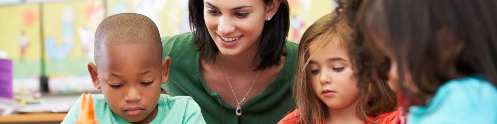 Teacher with short dark hair helps a diverse group of three young children in a colourful classroom. They focus on a shared activity
