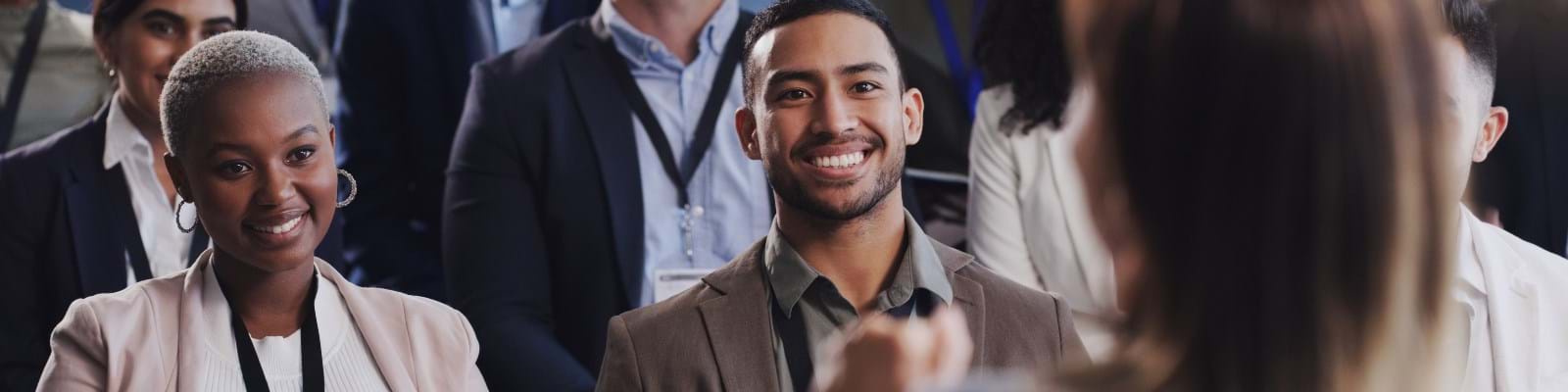 Audience members in business attire are smiling attentively at a speaker. 