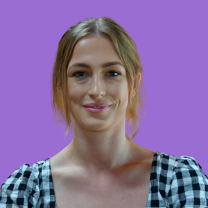 A woman with light brown hair smiles gently, wearing a black and white checkered top against a solid purple background.