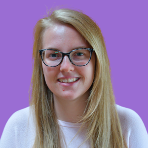 Smiling woman with long blonde hair and glasses set against a purple background. She wears a light pink sweater