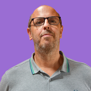 A middle-aged man with glasses and a beard stands against a purple background, wearing a grey polo shirt.
