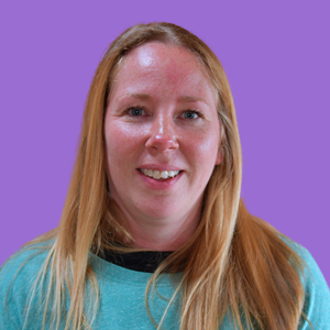 A woman with long red hair and a teal shirt smiles against a solid purple background.