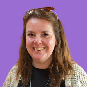 Smiling woman with long brown hair and sunglasses on head, against a solid purple background.