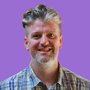 Smiling man with a greying beard and combed-back hair, wearing a checkered shirt. He stands against a solid purple background