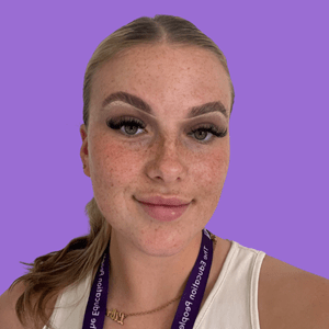 A young woman with freckles and hair in a low ponytail stands against a purple background. She wears a white top and a purple lanyard.