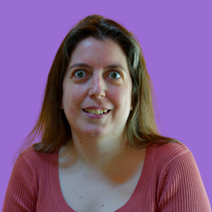 A woman with long brown hair and a pink top smiles at the camera against a solid purple background