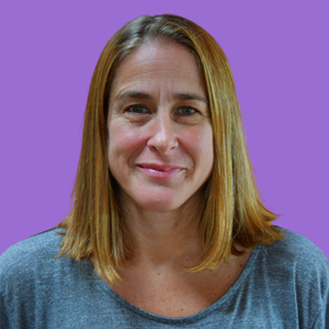 Woman with shoulder-length brown hair smiles gently against a solid purple background. She wears a grey top