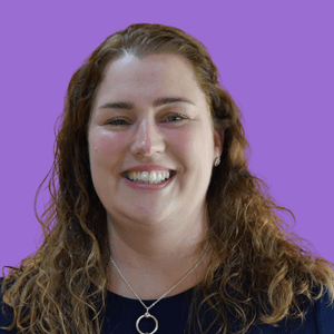 Smiling woman with curly hair against a purple background, wearing a silver necklace and earrings. 