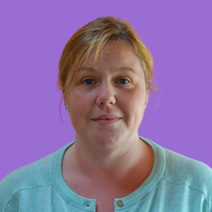 A woman with light brown hair and a slight smile wears a light blue top. She is set against a plain purple background
