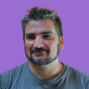A bearded man with short, spiky hair smiles subtly against a purple background. He wears a casual grey T-shirt